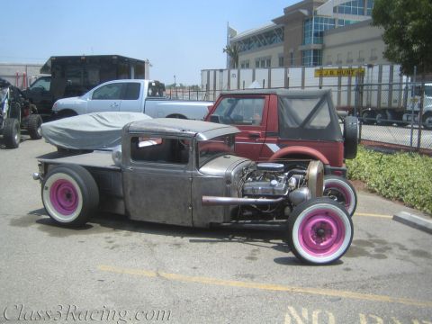 1990 Ford Bronco - low bare-metal rat-rod pickup with an exposed engine and pink whitewall wheels is parked in a lot in front of a covered car and red SUV.