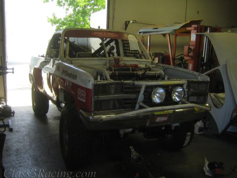 1990 Ford Bronco - white and red off-road race truck with a tubular front bumper and dual round lights sits being worked on inside a cluttered garage bay.