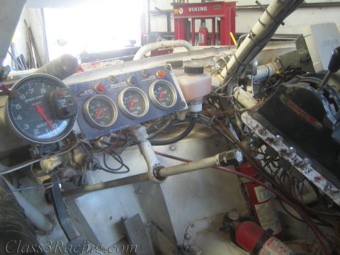 1990 Ford Bronco - cockpit of an off-road race truck shows a large tachometer, a row of auxiliary gauges and exposed roll-cage tubing inside a shop.