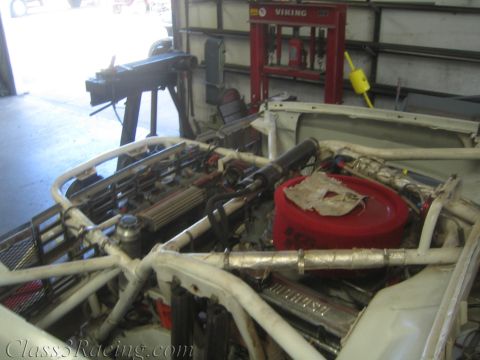 1990 Ford Bronco - open engine bay of a white off-road race vehicle shows a red round air cleaner, headers and surrounding roll-cage tubing inside a shop.