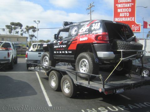 1990 Ford Bronco - black and red FJ Cruiser race truck numbered 348 sits on a flatbed trailer in a lot next to a 'Mexico Auto Insurance' billboard.