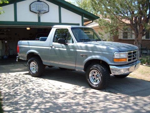 1994 Ford Bronco - Passenger-side front three-quarter view of a light blue full-size Ford Bronco with its removable hardtop off, parked on a driveway in front…
