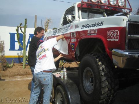1990 Ford Bronco - man in a printed T-shirt applies vinyl graphics to the fender of a red and white off-road race truck loaded with BFGoodrich and Bilstein sp…
