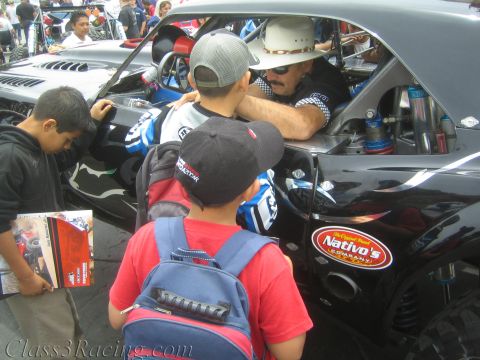 1990 Ford Bronco - Several children crowd around a black tube-frame race car while a man in a white cowboy hat leans out, near a Nativo's sponsor decal.