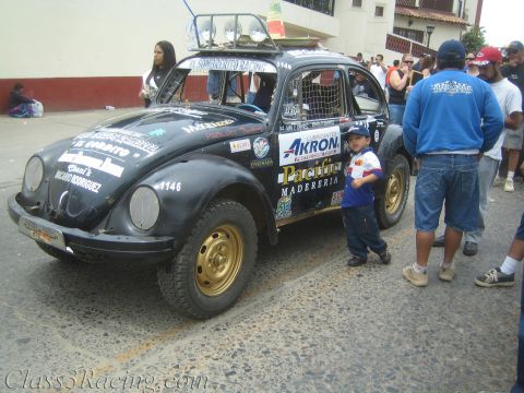 1990 Ford Bronco - black Baja-prepped Volkswagen Beetle race car covered in sponsor decals sits on a street, a small child standing beside it.