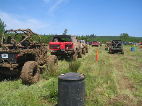 1989 Ford Bronco - Off-road vehicles are parked in a grassy field, with some covered in mud, under a clear blue sky.