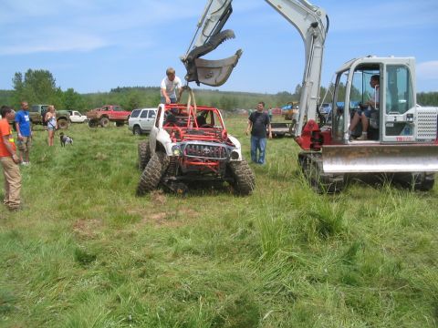 1989 Ford Bronco - Excavator lifts a heavily modified off-road vehicle in a grassy field with other vehicles present.