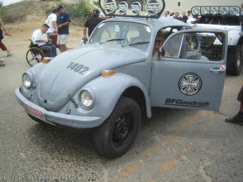 1990 Ford Bronco - primer-gray Baja-prepped Volkswagen Beetle numbered 1402 with a Hella roof light bar and BFGoodrich decals parked on a road.