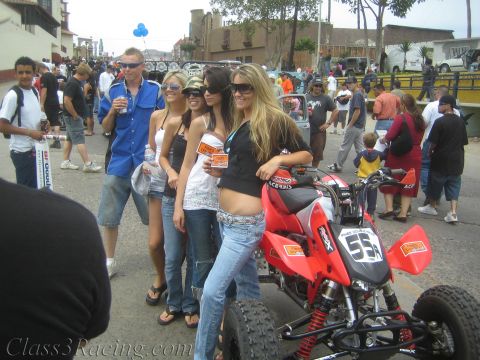 1990 Ford Bronco - Four young women pose beside a red number-55 racing ATV on a crowded street lined with spectators and chase trucks.