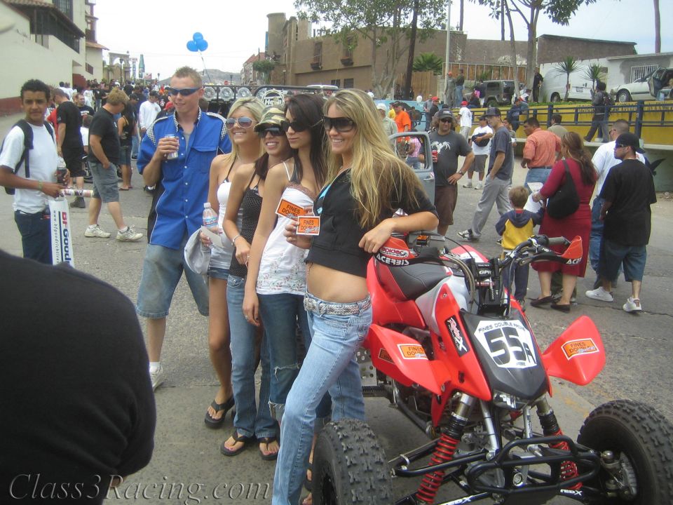 Four young women in casual clothes posing beside a red racing ATV in a street crowd