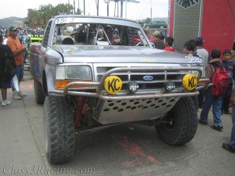 1990 Ford Bronco - silver Ford prerunner race truck numbered 1502 with yellow KC light pods and a tube bumper rolls through a street crowded with onlookers.