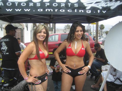 1990 Ford Bronco - Two women in matching red tops pose under a black AMRacing.tv promotional canopy at a street event, with a red pickup parked behind.