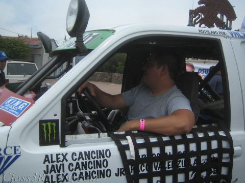 1990 Ford Bronco - man sits in the cab of a white off-road race truck with a Monster sticker and Alex Cancino lettering on the door.