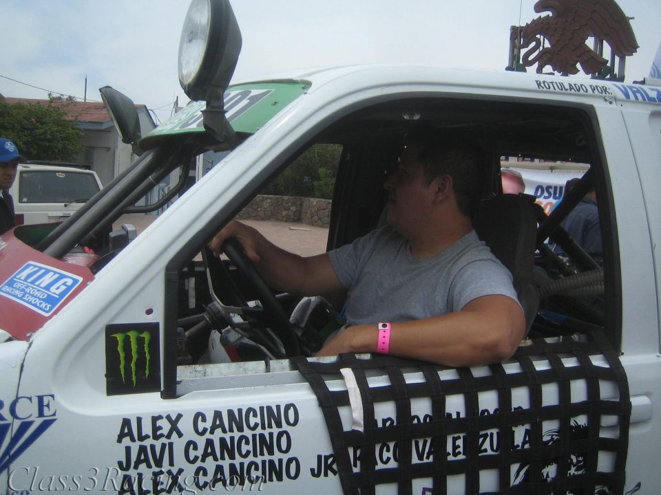 Driver seated in a white off-road race truck cab lettered Alex Cancino