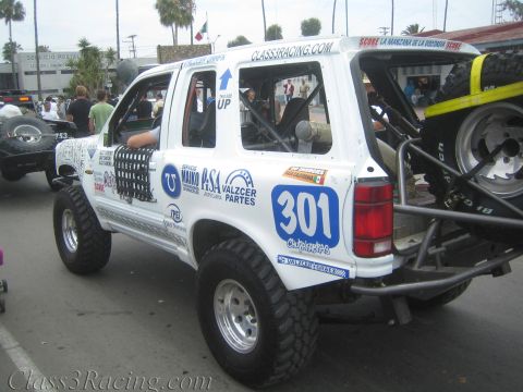 1990 Ford Bronco - lifted white Ford Explorer race truck numbered 301 with sponsor decals and a rear-mounted spare tire on a street with spectators.