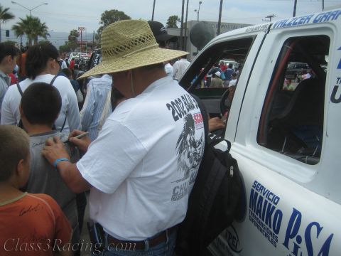 1990 Ford Bronco - man in a straw hat and a Baja-racing T-shirt stands among a crowd beside a white sponsor-lettered race vehicle.