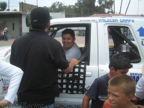 1990 Ford Bronco - smiling driver sits in the cab of a white Valzcer Grafx-lettered race truck while a man films him and children look on.