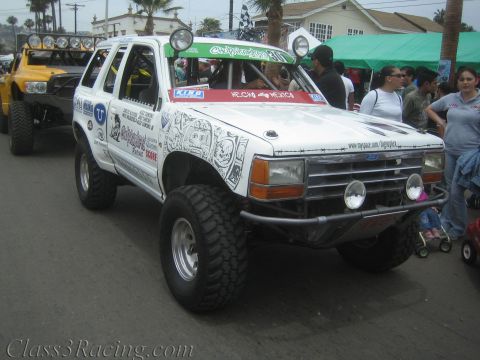 1990 Ford Bronco - lifted white Ford Explorer race truck with a roof light bar and graffiti-style hood art moves through a crowded street festival.