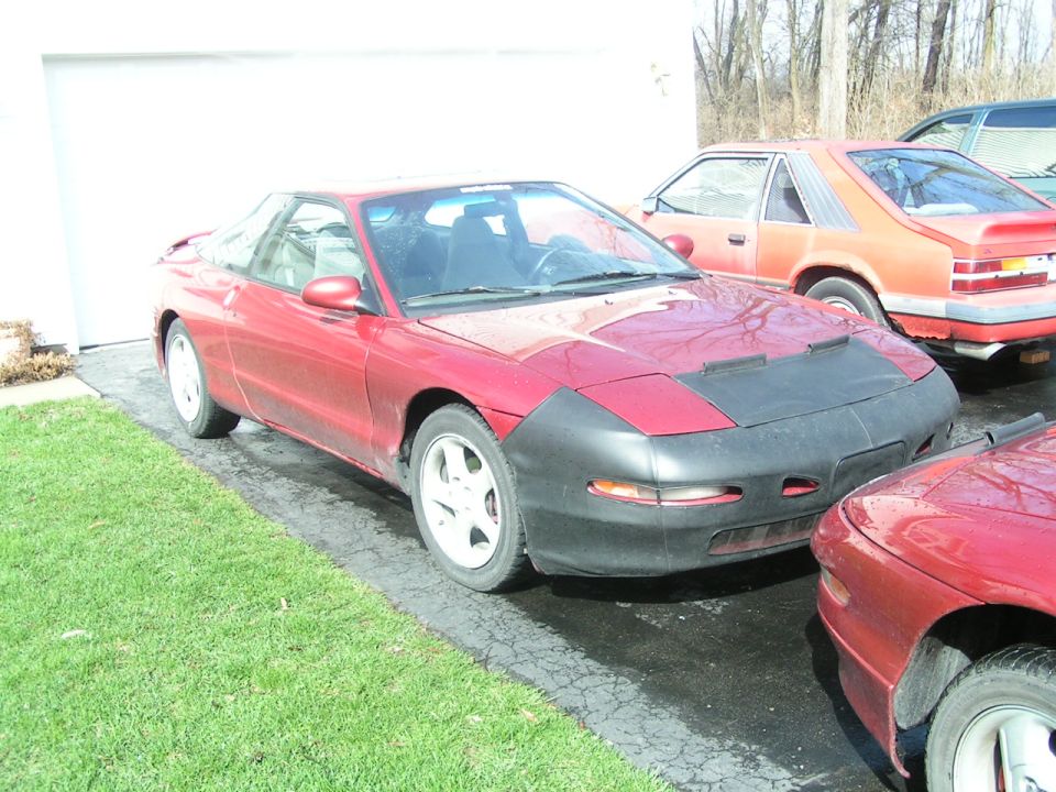 Red 1996 Ford Probe with a black car bra parked in a driveway next to another car
