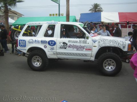 1990 Ford Bronco - lifted white Ford Explorer race truck numbered 301 shown in profile, covered in sponsor decals, amid a street crowd and tents.