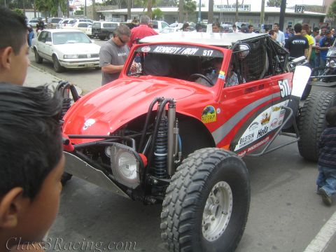 1990 Ford Bronco - red tube-frame off-road race buggy numbered 517 with Bilstein and Hella decals sits in a parking lot surrounded by spectators.