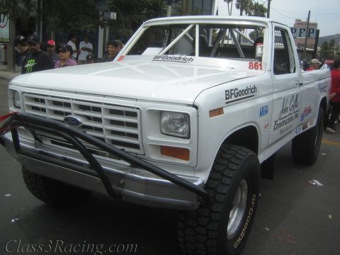 1990 Ford Bronco - white square-body Ford pickup race truck numbered 861 with BFGoodrich decals and a tube bumper moves through a street crowd.