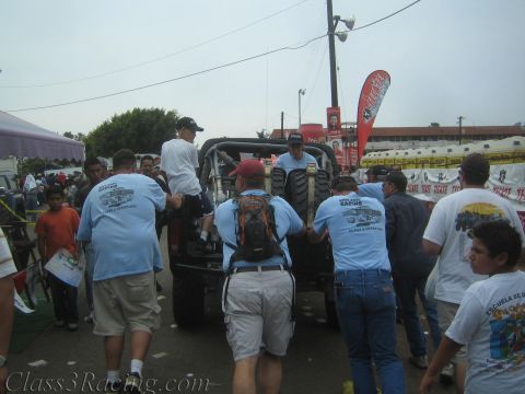 1990 Ford Bronco - crowd of men in racing-team T-shirts gather around the back of an off-road vehicle at an outdoor event with banners and tents.