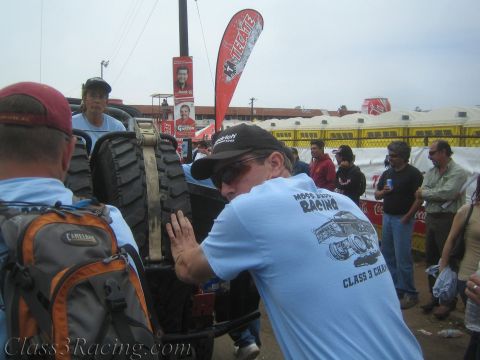 1990 Ford Bronco - man in a Class 3 racing-team T-shirt leans against a large knobby tire of an off-road vehicle in a busy outdoor event crowd.