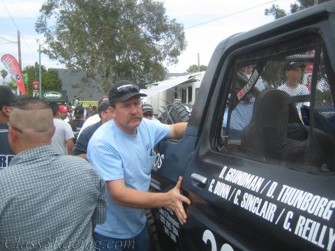 1990 Ford Bronco - man in a cap and blue shirt gestures beside the door of a black race vehicle lettered with driver names like D. Grundman and C. Reilly.