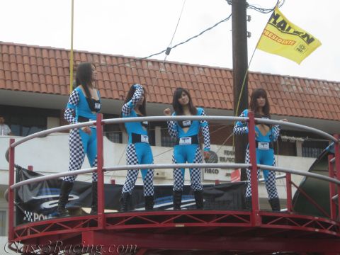 1990 Ford Bronco - Four women in matching blue-and-checkered racing suits pose on a red railed platform above a street with a yellow flag behind them.