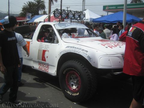 1990 Ford Bronco - white off-road race truck marked Rescue Racing sits among a crowd of spectators near a Pemex station, fitted with a roof light bar and spar…