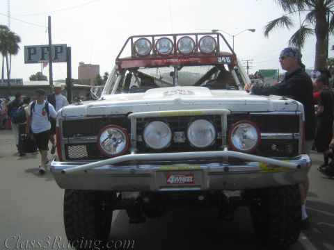 1990 Ford Bronco - Front view of a battered white off-road race truck with a roof light bar and round driving lights, parked on a street near a PFP sign.