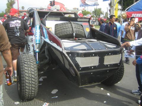 1990 Ford Bronco - Rear three-quarter view of a black tube-frame off-road race buggy with blue shocks and a spare tire, surrounded by spectators.