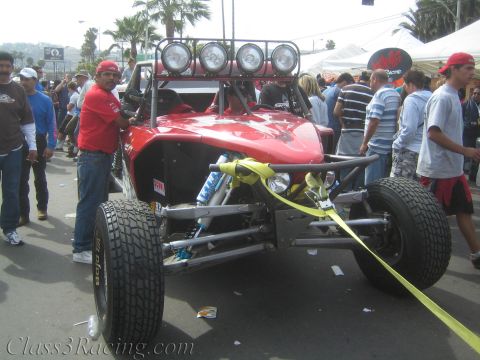 1990 Ford Bronco - red tube-frame off-road race buggy with a roof light bar is towed by a yellow strap through a crowd at an outdoor event.