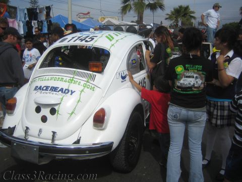 1990 Ford Bronco - Rear view of a white off-road racing Volkswagen Beetle marked 1145 and Villanos Racing, with people gathered around it on a street.