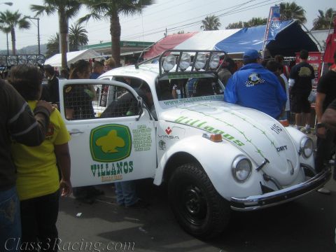 1990 Ford Bronco - white off-road racing Volkswagen Beetle with Villanos graphics, a roll cage and roof light bar parked at a busy event area.