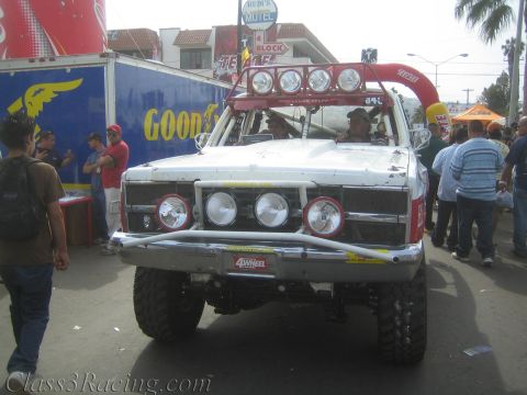 1990 Ford Bronco - Front view of a white off-road race truck with roof light bar parked on a crowded street near a Rudi's Motel sign and red tents.