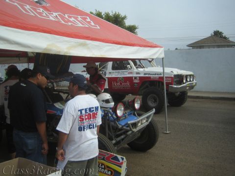 1990 Ford Bronco - People gather under a red tent beside a white off-road race truck and a blue tube-frame buggy at a tech inspection area.