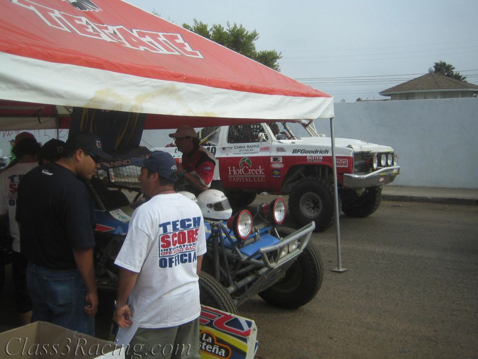 Off-road race vehicles parked under a red tent with people gathered around