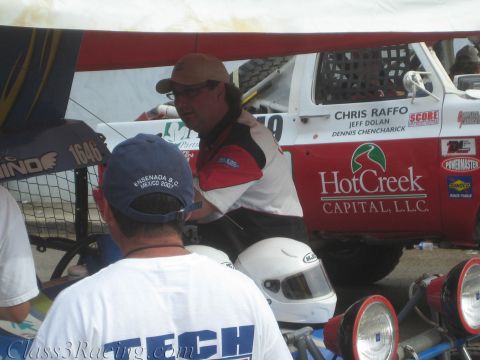 1990 Ford Bronco - man in a tan cap leans by a white race car door lettered Chris Raffo, Jeff Dolan and HotCreek Capital, with helmets in the foreground.