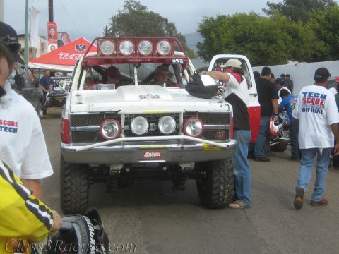 1990 Ford Bronco - white off-road race truck with roof light bar moves through a tech inspection lane flanked by officials in Score Tech shirts.