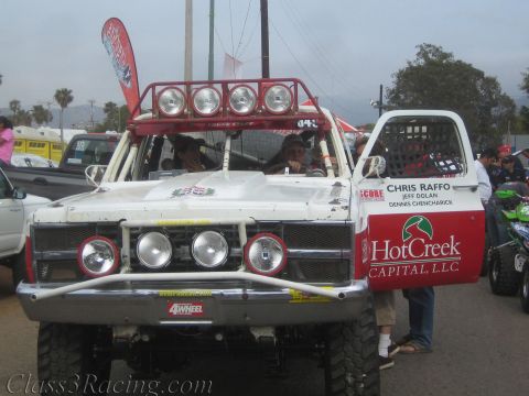 1990 Ford Bronco - Front three-quarter view of a white off-road race truck with roof light bar, lettered Chris Raffo and HotCreek Capital on the open door.