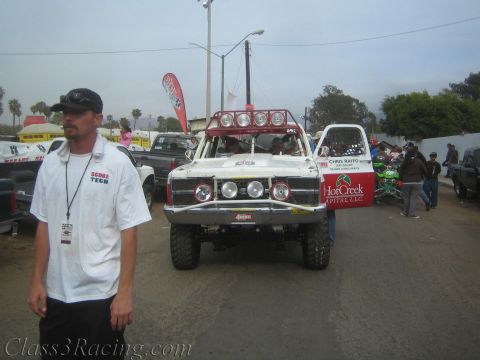1990 Ford Bronco - Score Tech official in a white shirt walks past a white off-road race truck with HotCreek Capital lettering on a gray overcast day.