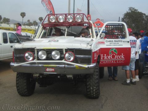 1990 Ford Bronco - Front view of a white off-road race truck with roof light bar and HotCreek Capital door lettering, with people standing alongside.
