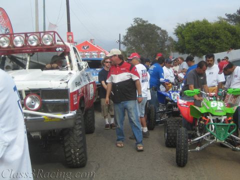 1990 Ford Bronco - crowd mingles on a street between a white off-road race truck on the left and green and blue racing ATVs on the right.