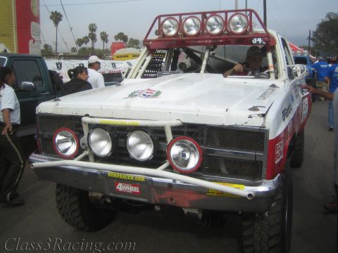 1990 Ford Bronco - Front view of a white off-road race truck with roof light bar, round driving lights and patriotic crest, parked among palm trees.
