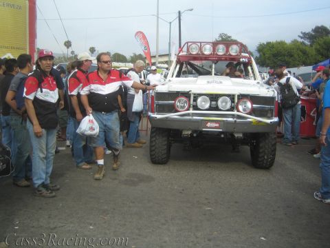 1990 Ford Bronco - white off-road race truck with roof light bar rolls through a crowd that includes men in red and black team shirts at an event.