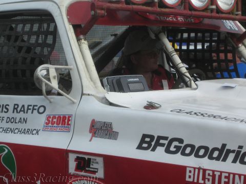 1990 Ford Bronco - driver in a cap sits in the caged cab of a white off-road race truck lettered with Raffo names, BFGoodrich, Bilstein and Score.