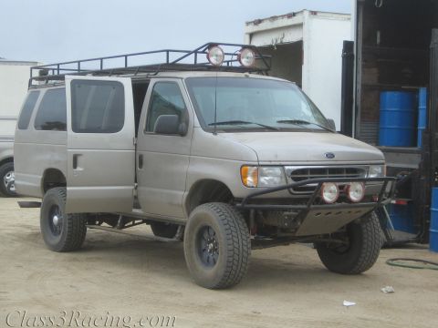 1990 Ford Bronco - silver Ford full-size van lifted on off-road tires with a roof rack, auxiliary lights and tube front bumper parked on a dirt lot.