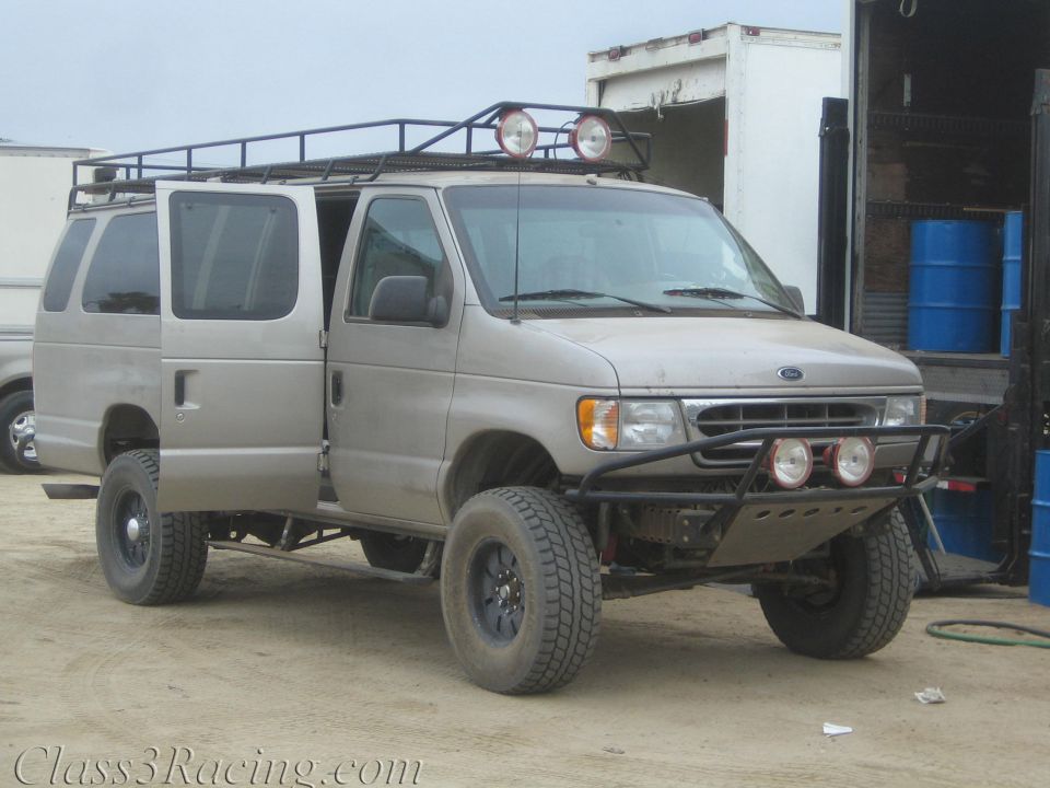 Silver Ford full-size van with roof rack, off-road bumper and lift on dirt lot